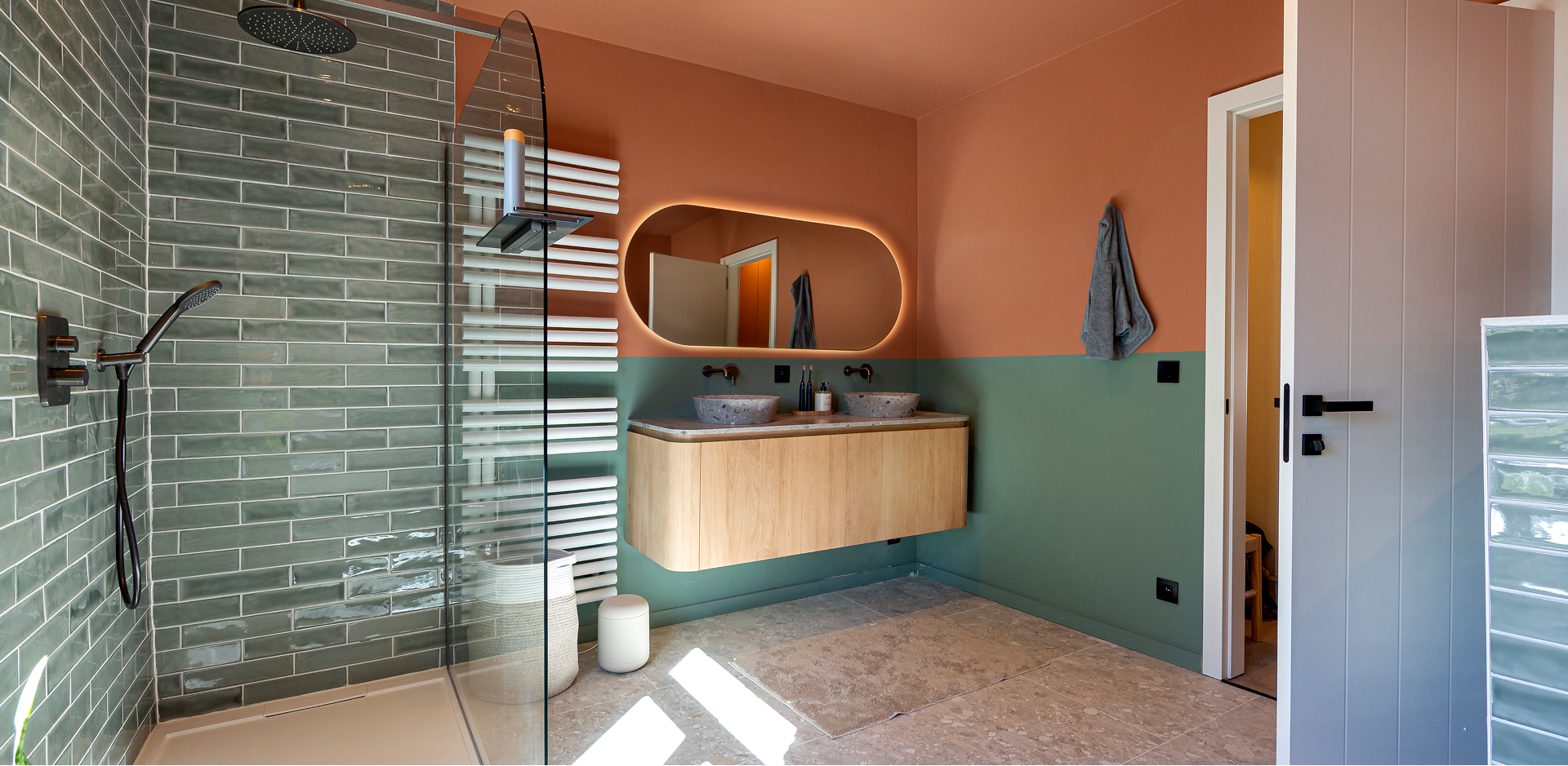 Moderne badkamer met groene tegelwand, inloopdouche, houten dubbel wastafelmeubel, verlichte organische spiegel en muren in terracotta en groen.