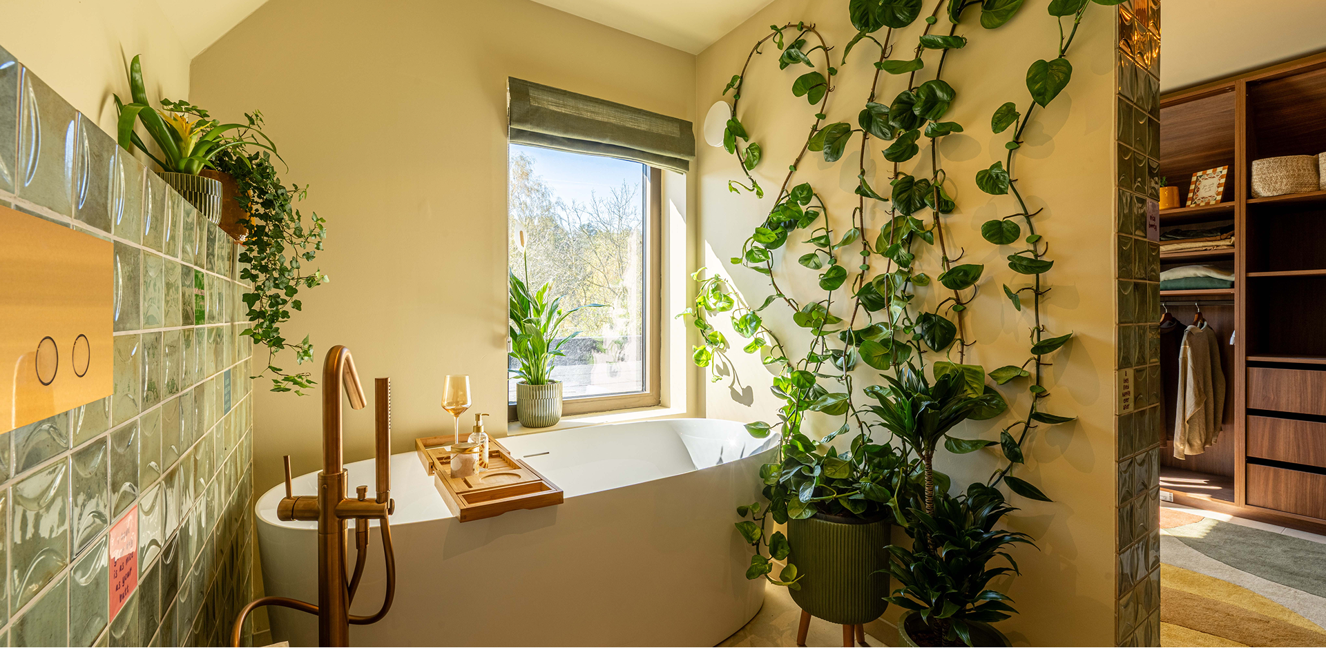 Badkamer met vrijstaand bad, groene glastegels, gouden kraanwerk, veel kamerplanten en een open dressing naast de badruimte.