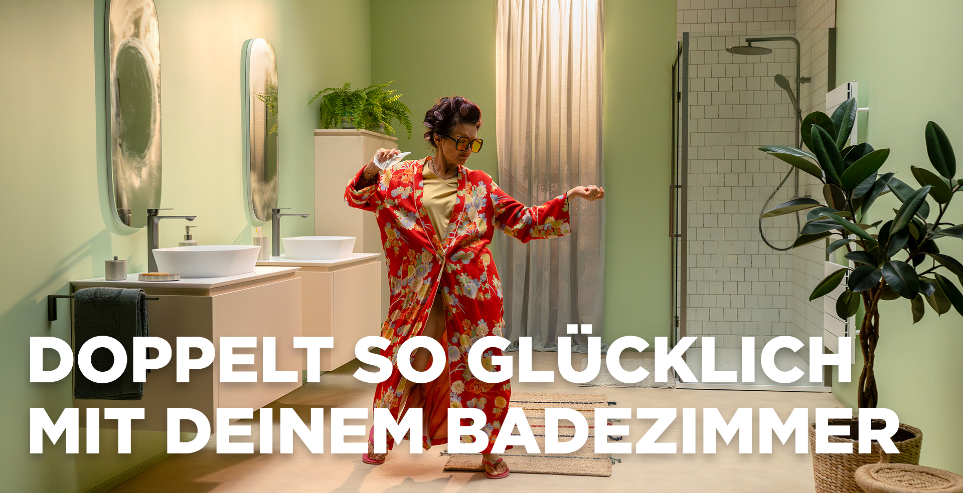 In dieser lebendigen Szene von x²o Badezimmer siehst du eine fröhliche Frau, die in einem modernen Badezimmer mit hellgrünen Wänden tanzt. Sie trägt einen roten Bademantel mit Blumenmuster und steht zwischen einem Doppelwaschtisch mit zwei Waschbecken und Spiegeln auf der linken Seite und einer begehbaren Dusche mit Glaswand und weißen Fliesen auf der rechten Seite. Pflanzen und eine Badematte sorgen für eine warme Atmosphäre.