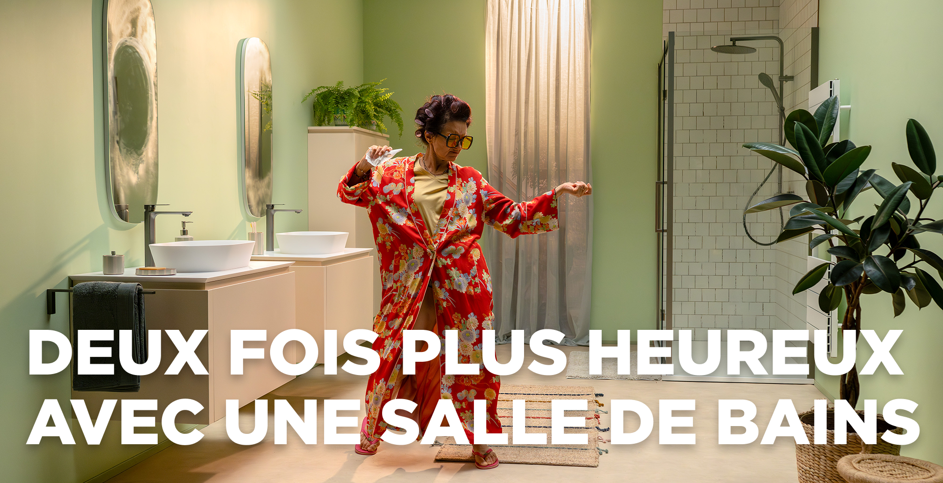 Dans cette scène dynamique de x²o salles de bains, une femme joyeuse danse dans une salle de bains moderne aux murs vert clair. Elle porte un peignoir rouge à fleurs et se tient entre un meuble double vasque avec deux lavabos et miroirs à gauche et une douche à l’italienne avec paroi en verre et carrelage blanc à droite. Des plantes et un tapis de bain apportent une ambiance chaleureuse.