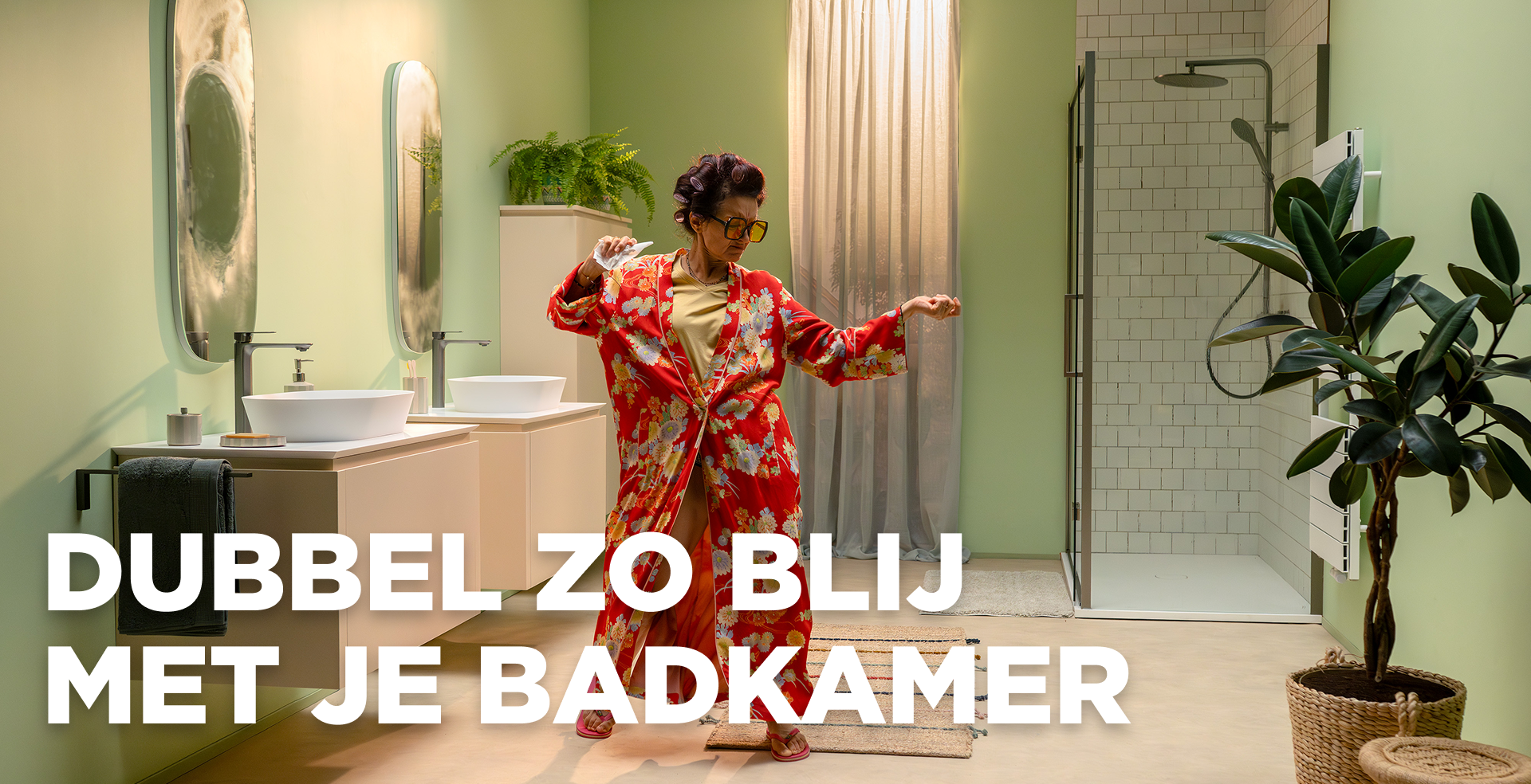 In deze levendige scène van x²o badkamers zie je een vrolijke vrouw die danst in een moderne badkamer met lichtgroene muren. Ze draagt een rode kamerjas met bloemenprint en staat tussen een dubbel wastafelmeubel met twee waskommen en spiegels aan de linkerkant en een inloopdouche met glazen wand en witte tegels aan de rechterkant. Er zijn planten en een zachte badmat aanwezig die de ruimte een warme uitstraling geven. Op de afbeelding staat de tekst: “DUBBEL ZO BLIJ MET JE BADKAMER”.