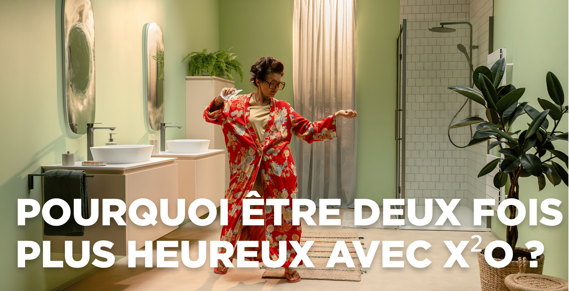 Dans cette scène dynamique de x²o salles de bains, une femme joyeuse danse dans une salle de bains moderne aux murs vert clair. Elle porte un peignoir rouge à fleurs et se tient entre un meuble double vasque avec deux lavabos et miroirs à gauche et une douche à l’italienne avec paroi en verre et carrelage blanc à droite. Des plantes et un tapis de bain apportent une ambiance chaleureuse.