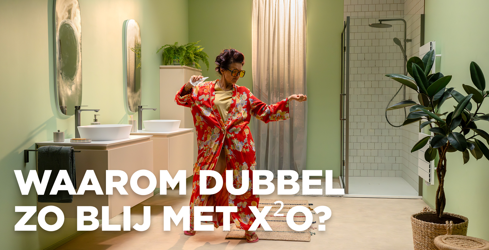 In deze levendige scène van x²o badkamers zie je een vrolijke vrouw die danst in een moderne badkamer met lichtgroene muren. Ze draagt een rode kamerjas met bloemenprint en staat tussen een dubbel wastafelmeubel met twee waskommen en spiegels aan de linkerkant en een inloopdouche met glazen wand en witte tegels aan de rechterkant. Er zijn planten en een zachte badmat aanwezig die de ruimte een warme uitstraling geven. 