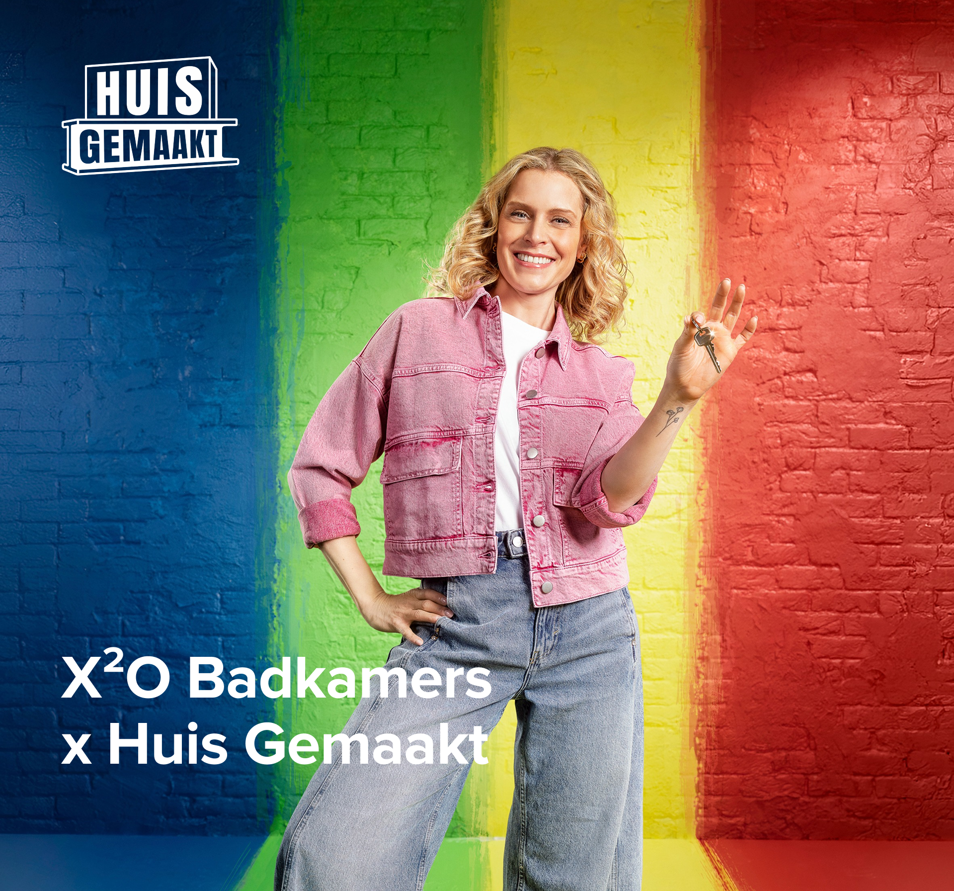Banner van X²O badkamers in samenwerking met Huis Gemaakt, met een glimlachende vrouw in een roze jas die een sleutel vasthoudt voor een bakstenen muur in blauw, groen, geel en rood, met het logo “Huis Gemaakt” links in beeld.