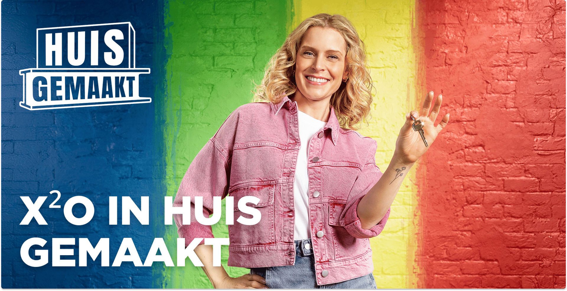 Vrouw met krullend blond haar in roze jeansjasje houdt sleutels omhoog en glimlacht voor een bakstenen muur in regenboogkleuren met de tekst “Huis Gemaakt” en “X²O in Huis Gemaakt”, campagnebeeld van x²o badkamers.