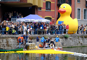 Mensen staan langs een kade bij het water terwijl enkele personen tussen drijvende gele badeendjes in het kanaal staan; op de achtergrond staat een grote opblaasbare badeend en een tent van X²O badkamers.