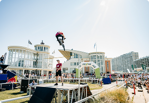 Foto van een event op het strand met een podium, meerdere mensen, gebouwen op de achtergrond en een persoon die hoog in de lucht boven het terrein hangt, met zichtbare aanwezigheid van X²O badkamers op het evenement.