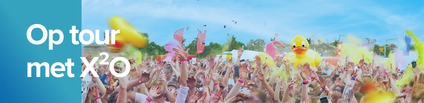 Een uitbundige festivalmenigte onder een blauwe lucht, gooit kleurpoeder in de lucht met een grote opblaasbare badeend in het midden, tijdens de zomertour van X²O badkamers.