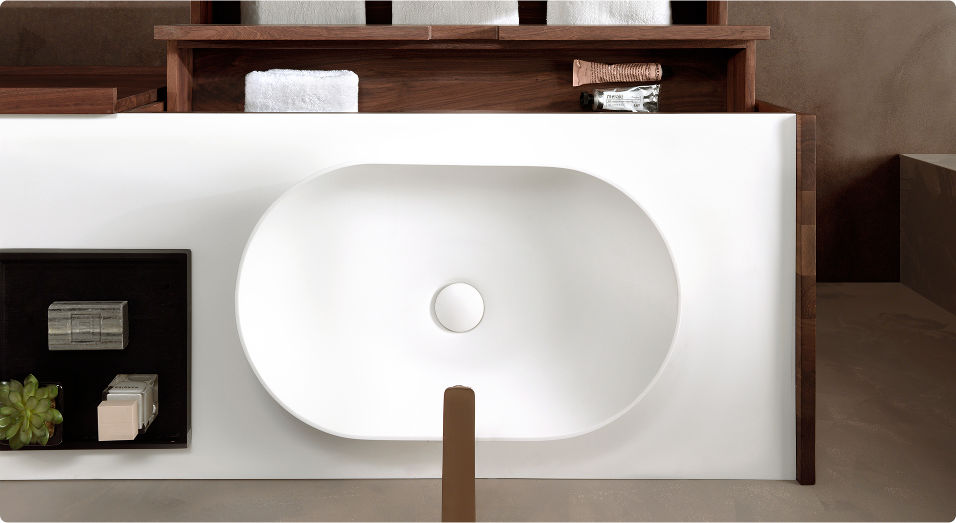 Bovenaanzicht van een minimalistische, witte wasbak ingebouwd in een meubel met houten accenten, onderdeel van een x²o badkamers interieur. Links op het werkblad staat een zwarte tray met decoratieve zepen en een klein vetplantje. Op de achtergrond zijn houten open vakken met handdoeken en verzorgingsproducten zichtbaar.