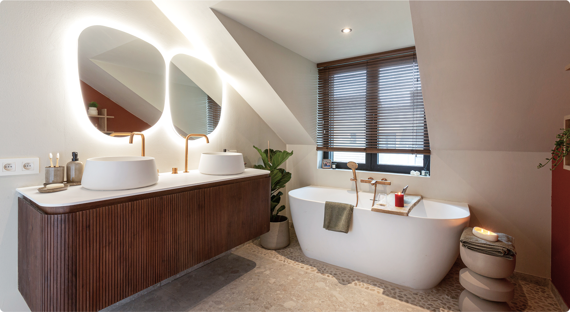 Moderne x²o badkamers inrichting onder een schuin dak met dubbele ronde spiegels, sfeerverlichting, twee vrijstaande waskommen met koperkleurige kranen, een vrijstaand bad voor het raam met houten jaloezieën en accessoires zoals kaarsen en planten.