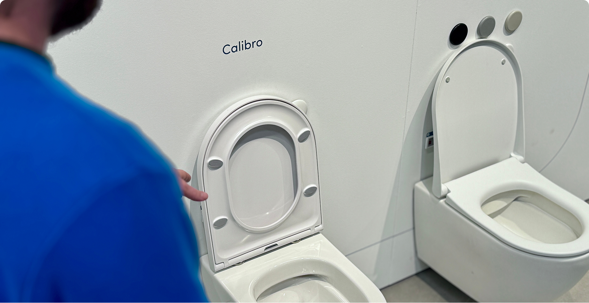 Een verkoper in blauwe trui wijst naar een toiletzitting met de merknaam Calibro in een toonzaal. Advies op maat bij x²o badkamers.