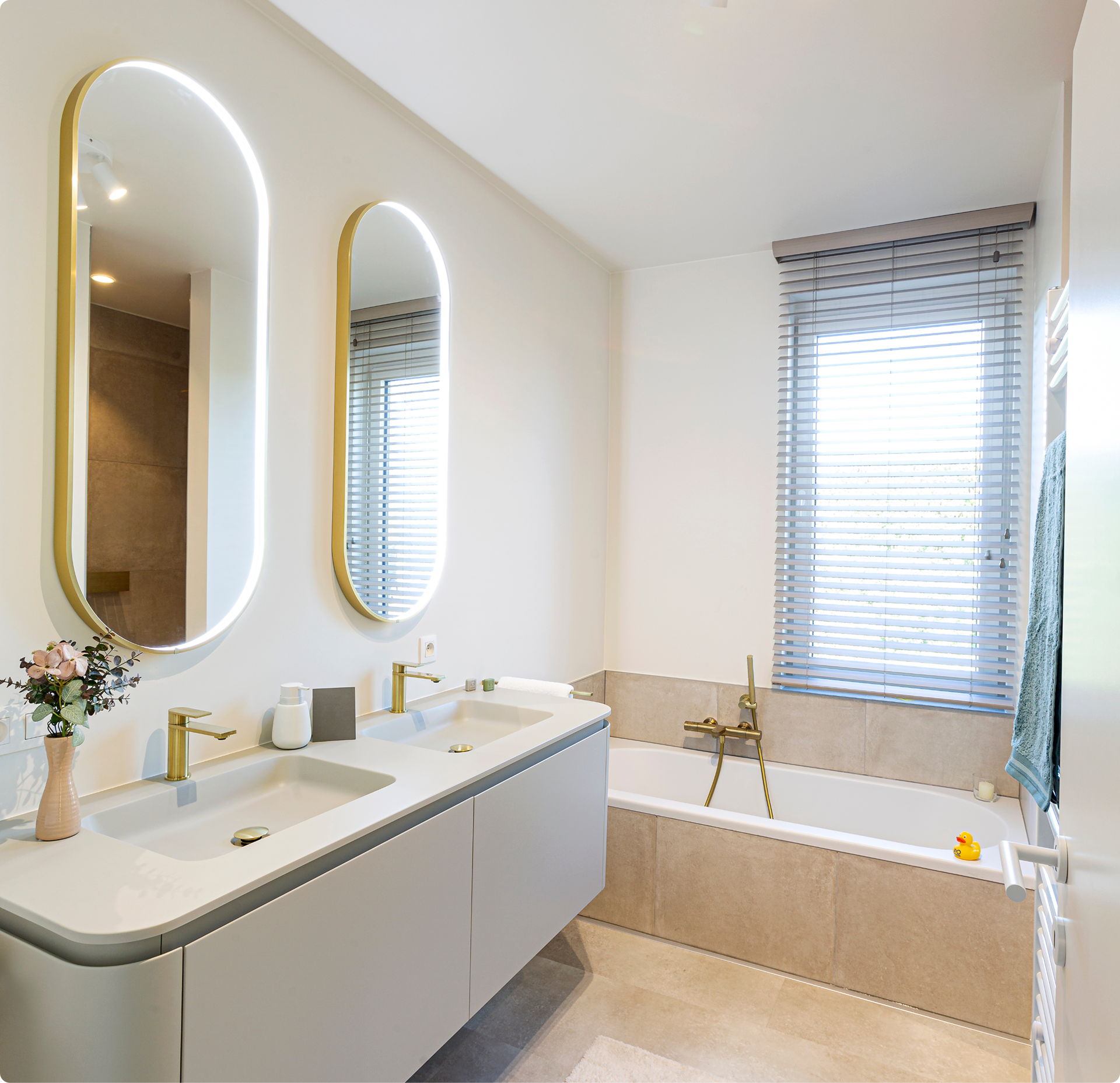 Lichte, frisse x²o badkamers met moderne indeling. Links een zwevend meubel met dubbele wastafel, goudkleurige kranen en twee verticale spiegels met ledverlichting. Rechts een ingebouwd ligbad met bijpassende kraan en een raam met jaloezieën zorgt voor natuurlijk licht.