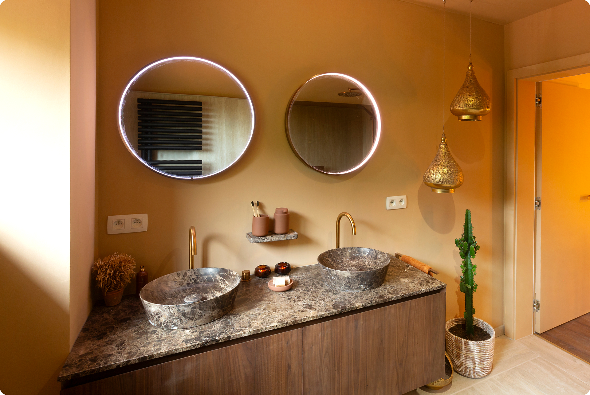 Warme, oosterse sfeer in deze x²o badkamers met aardetinten, twee ronde spiegels met geïntegreerde verlichting en een dubbel wastafelmeubel in hout met marmerlook werkblad. Op de achtergrond hangen decoratieve lantaarns en een cactus staat in een mand links van de deuropening.