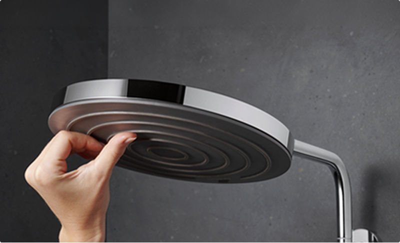 Close-up van een ronde douchekop van x²o badkamers waarbij een hand de sproeikoppen aanraakt, zichtbaar tegen een donkere achtergrond.