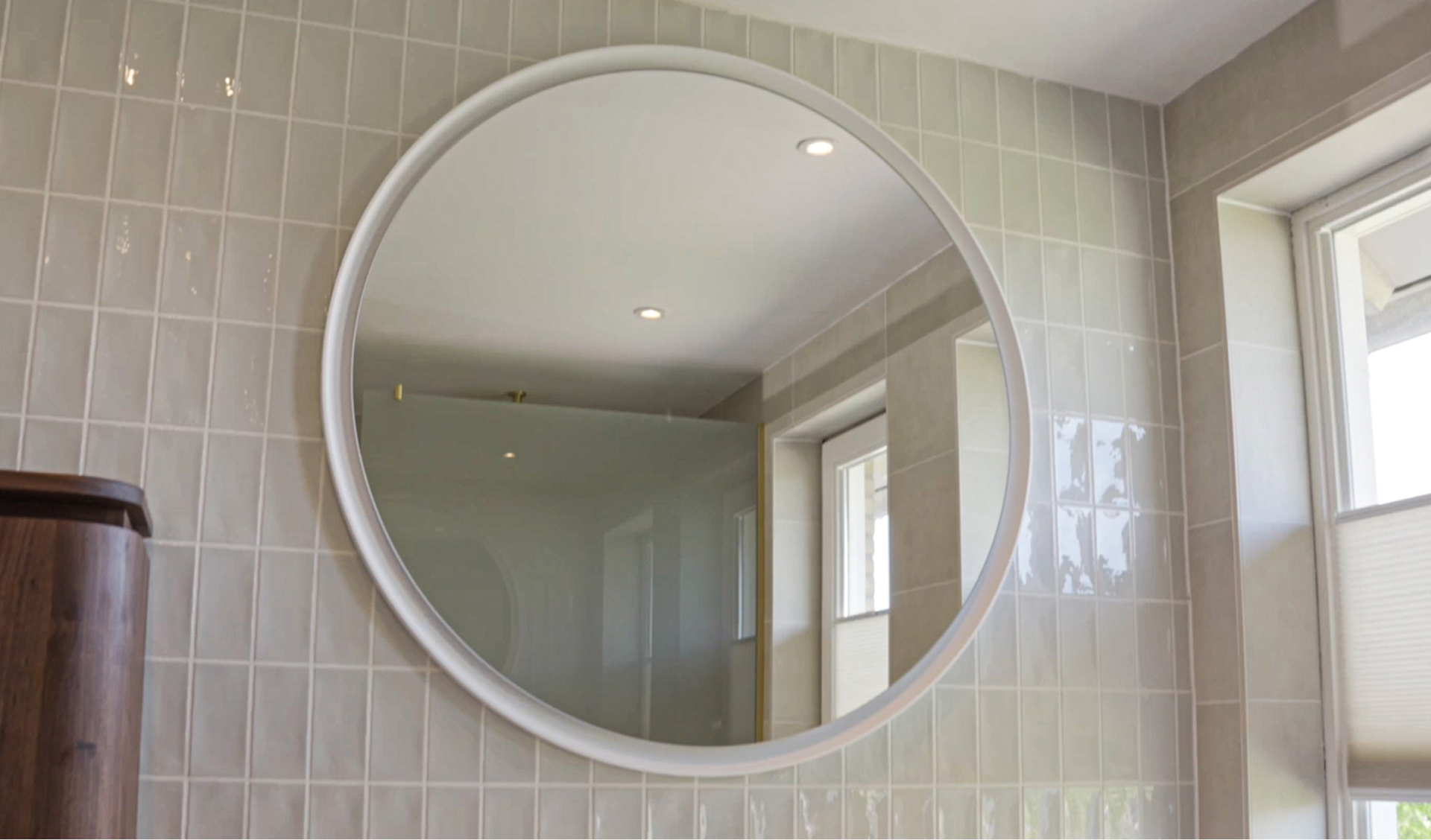 Runder Spiegel mit weißem Rahmen an einer Wand mit glänzenden rechteckigen Fliesen. Der Spiegel reflektiert Tageslicht und Einbauspots. x²o Bad.