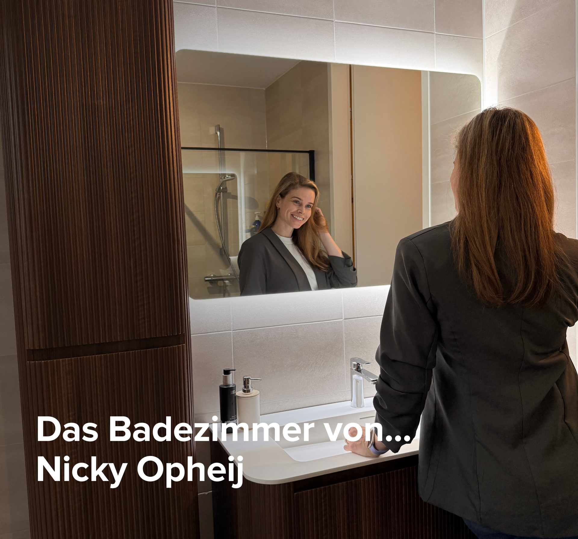 In dieser inspirierenden Szene von x²o Badezimmer siehst du ein stilvolles Badezimmer mit einem Holzmöbel und einem beleuchteten Spiegel. Eine Frau lächelt und betrachtet sich im Spiegel, während sie ihr Haar berührt. Auf dem Bild steht der Text: „De badkamer van… Nicky Opheij“.