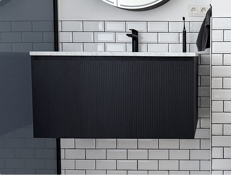 Zwevend badkamermeubel van x²o badkamers in zwart met verticale ribstructuur, geplaatst tegen een wand met witte metrotegels en zwarte leidingen.