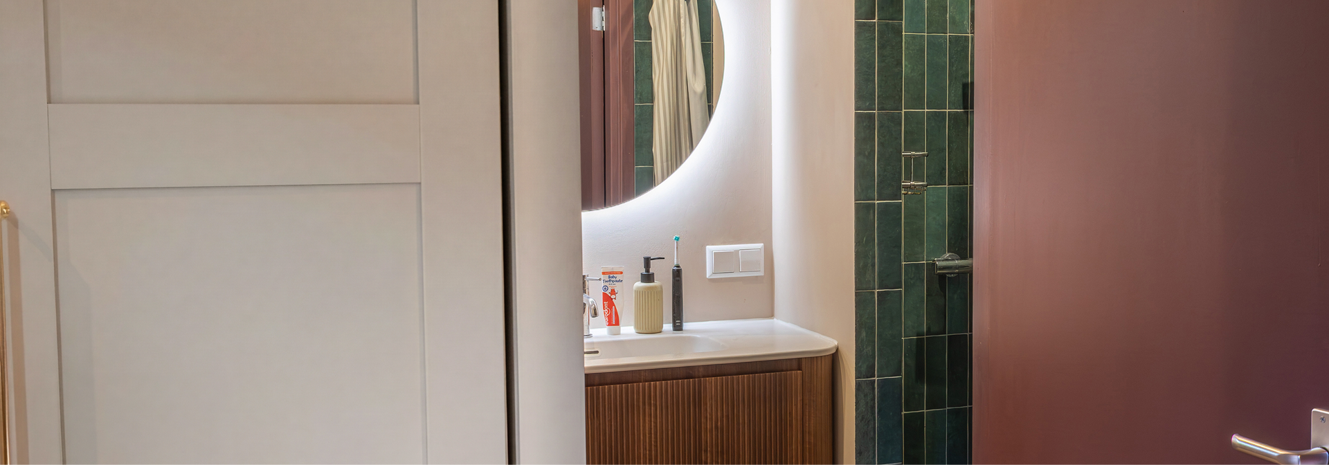 Gedeeltelijk zicht op een badkamer x²o badkamers met een halfopen deur die uitkijkt op een houten geribbeld wastafelmeubel met witte waskom, chromen kraan, ronde verlichte spiegel en rechts een douche met groene tegels en glazen wand