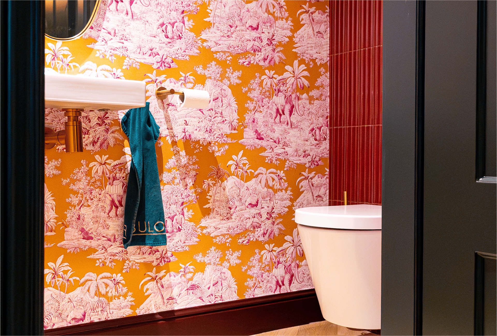 Goldener Toilettenpapierhalter im Stil von x²o Badezimmer mit zwei Rollen, vor roten Wandfliesen und rosafarbener Tapete.