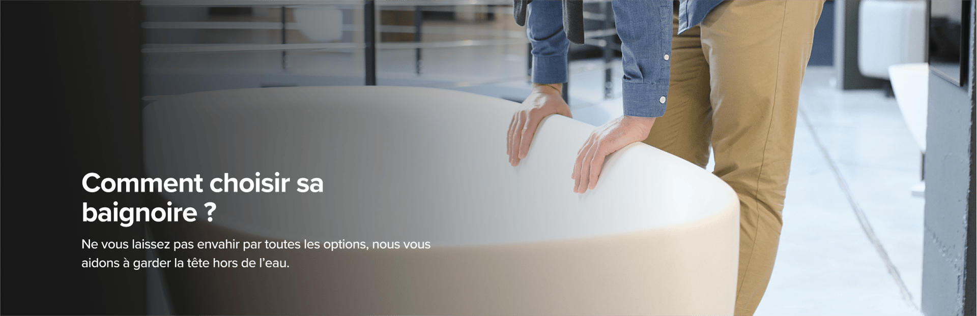 Sur cette image de x²o badkamers, un homme en pantalon beige et chemise bleue examine une grande baignoire design van x²o badkamers in een showroom met een industriële vloer en andere badmodellen op de achtergrond.