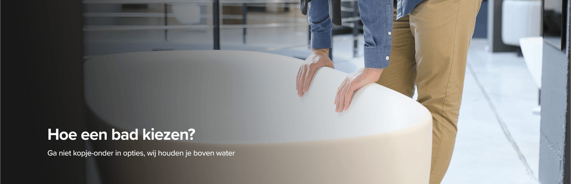 Op deze foto van x²o badkamers zie je een man in beige broek en blauwe blouse die een grote designbadkuip van x²o badkamers bekijkt in een showroom met een industriële vloer en andere badmodellen op de achtergrond.