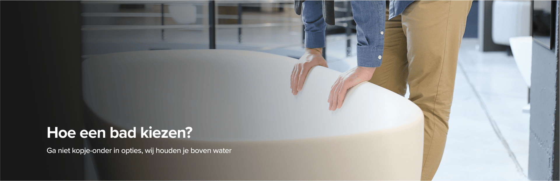 Op deze foto van x²o badkamers zie je een man in beige broek en blauwe blouse die een grote designbadkuip van x²o badkamers bekijkt in een showroom met een industriële vloer en andere badmodellen op de achtergrond.