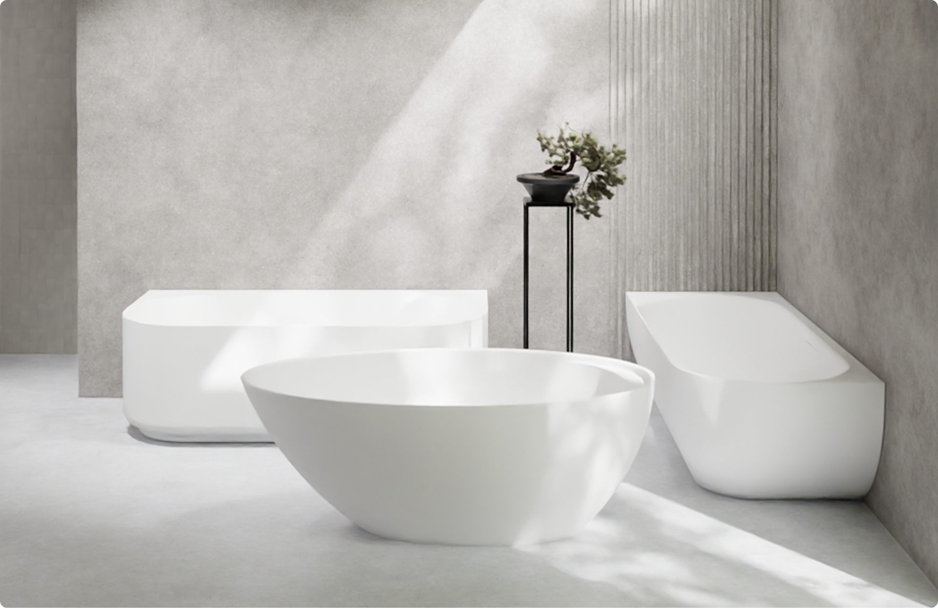 Op deze foto zie je drie verschillende vrijstaande baden in een minimalistische x²o badkamers setting. Vooraan staat een rond wit bad met zachte lijnen, links een rechthoekig bad met strakke hoeken en rechts een ovaal bad met een schuine rugleuning. De baden staan op een lichte betonnen vloer tegen grijze wanden met een subtiel verticaal reliëf. Een zwarte hoge plantenstandaard met een klein boompje maakt het serene design compleet.