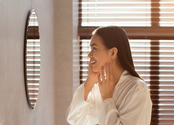 Eine lächelnde Frau im weißen Morgenmantel betrachtet sich im Wandspiegel eines hellen X²O Badezimmers mit Holzjalousien.