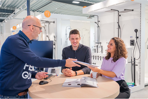 Een x²o badkamers showroom waar een medewerker een brochure overhandigt aan een glimlachende vrouw aan een ronde tafel. Een man zit ernaast, luisterend. Op de achtergrond staan moderne doucheopstellingen uitgestald.
