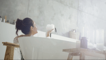 Une jeune femme se détend dans une baignoire blanche moderne et souffle de la mousse dans l’air de façon ludique.