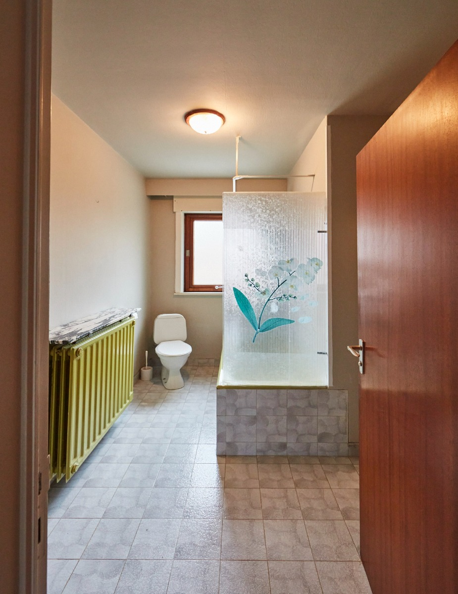 Veraltetes Badezimmer mit gelbem Heizkörper und Glas-Duschwand mit Blumenmotiv.