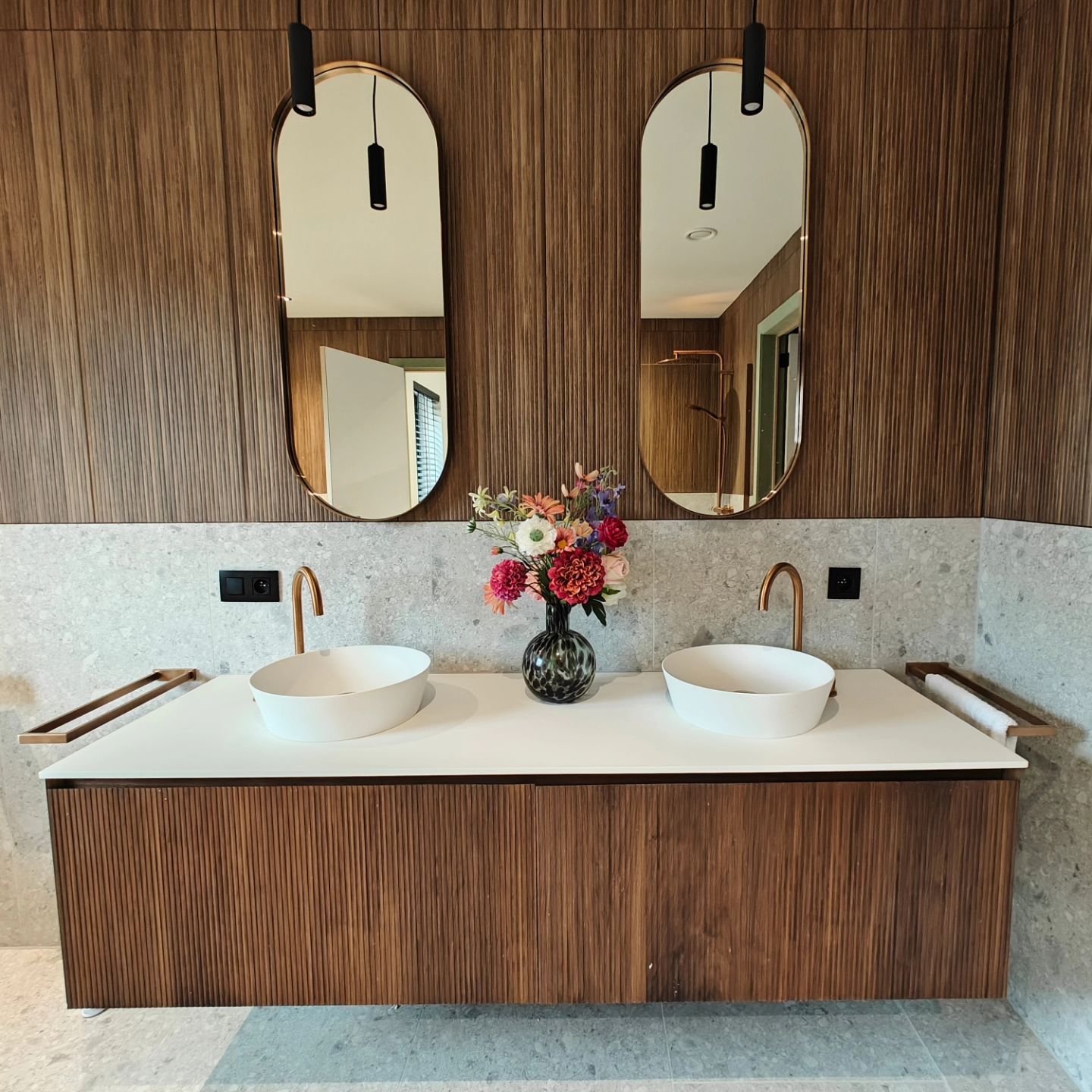 Badmöbel aus warmem Holz mit vertikaler Rillenstruktur und einer langen weißen Waschtischplatte mit zwei runden Aufsatzwaschbecken. Darüber hängen zwei abgerundete Spiegel mit schwarzen Pendelleuchten, in der Mitte steht eine Vase mit bunten Blumen; seitlich sind Handtuchhalter montiert und die Rückwand ist mit einer hellgrauen Steinoptik samt schwarzen Steckdosen verkleidet.