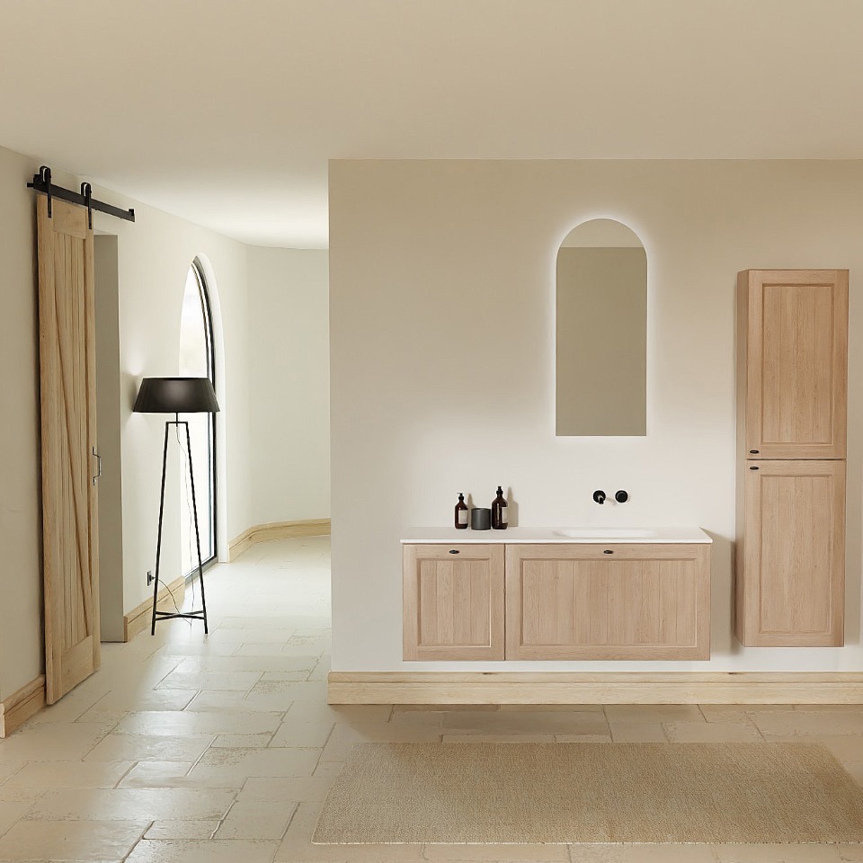 Ruhiges, minimalistisches Badezimmer mit einem schwebenden Badmöbel aus hellem Eichenholz, einem weißen Waschtisch und schwarzen Wandarmaturen. Über dem Möbel hängt ein bogenförmiger Spiegel mit dezenter LED-Hinterleuchtung. Rechts befindet sich ein passender Hochschrank aus hellem Holz. Links ist eine Holzschiebetür mit schwarzer Schiene sowie eine hohe schwarze Stehleuchte neben einer abgerundeten Fensteröffnung zu sehen. Der Boden besteht aus hellen Natursteinfliesen, vor dem Möbel liegt eine weiche Badematte.