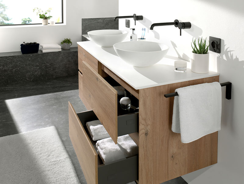 Stijlvolle badkamermeubelcompositie met houten wastafelmeubel, open lade met opgevouwen handdoeken, dubbele opbouwwaskommen en zwarte wandkranen, badend in natuurlijk daglicht.