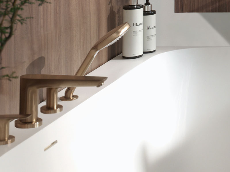 Moderne badkamerdetail met warme koperkleurige badrandkranen en een handdouche gemonteerd op een wit vrijstaand bad, afgewerkt met natuurlijke houtaccenten en zachte verlichting die een warme en luxueuze sfeer creëren.