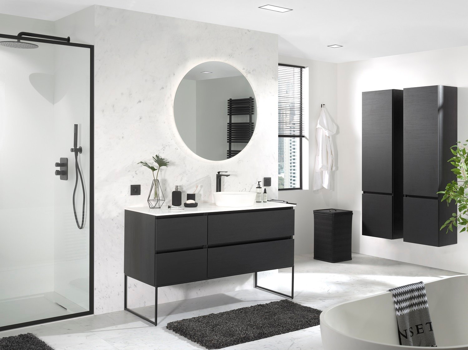 Moderne Komplettbadlösung mit schwarzem Holzwaschtisch auf schlankem Metallgestell, weißem Aufsatzwaschbecken und rundem Spiegel mit indirekter LED-Beleuchtung vor einer Wand in Marmoroptik. Links ist eine bodengleiche Dusche mit schwarzem Aluminiumprofil und Regendusche zu sehen, rechts ergänzen hohe schwarze Schränke, ein Fenster mit Jalousie und eine freistehende Badewanne das elegante Gesamtbild.