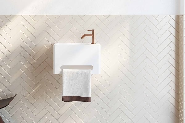 Kompaktes weißes Waschbecken mit integrierter Handtuchhalterung und kupferfarbenem Wasserhahn vor Fischgrätfliesen.