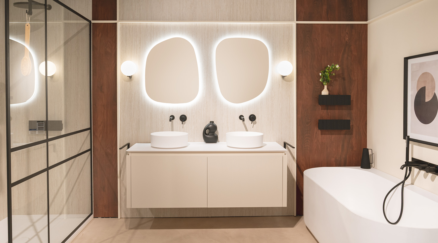 Moderne badkameropstelling van x²o badkamers met een zwevend crème badkamermeubel, twee ronde opzetwaskommen in mat wit en twee organisch gevormde spiegels met indirecte LED-verlichting. De achterwand bestaat uit lichte textuurpanelen gecombineerd met warme houten wandpanelen. Links is een zwarte industriële douchewand zichtbaar en rechts een vrijstaand wit bad met zwarte kraan.