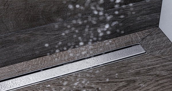 Nahaufnahme eines Duschabflusses mit einem Edelstahlkanal und Wassertropfen, die aus dem Duschkopf fließen, auf einem Holzoptik-Boden