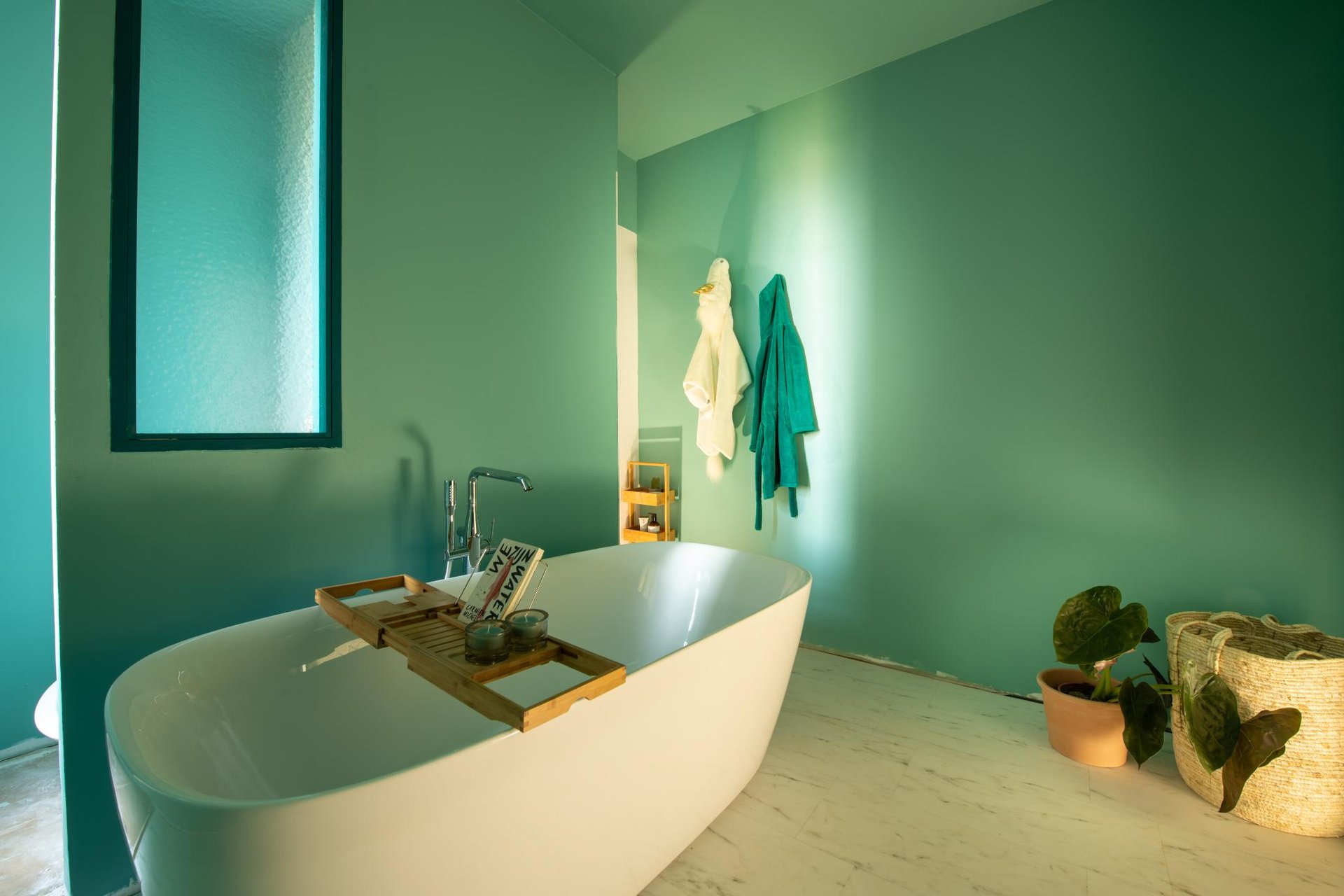 Badezimmer mit türkisfarbenen Wänden, freistehende Badewanne, Holzregal mit Accessoires, Handtücher an der Wand und Pflanzen auf dem Boden