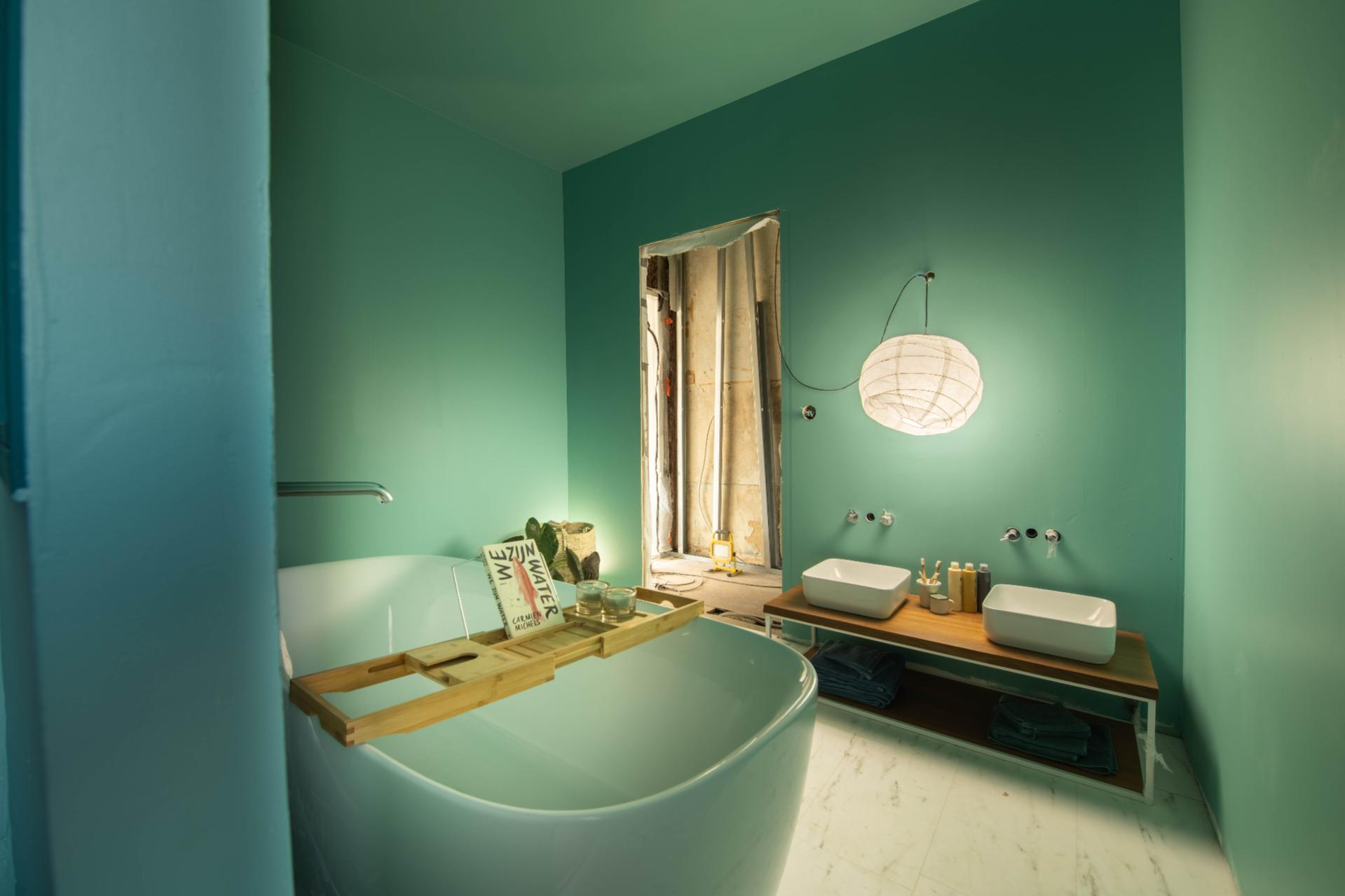 Stilvolles Badezimmer mit grünen Wänden, freistehender Badewanne mit Holzregal, zwei Waschbecken und warmem Licht. Das Fenster ist mit einem Stoff bedeckt