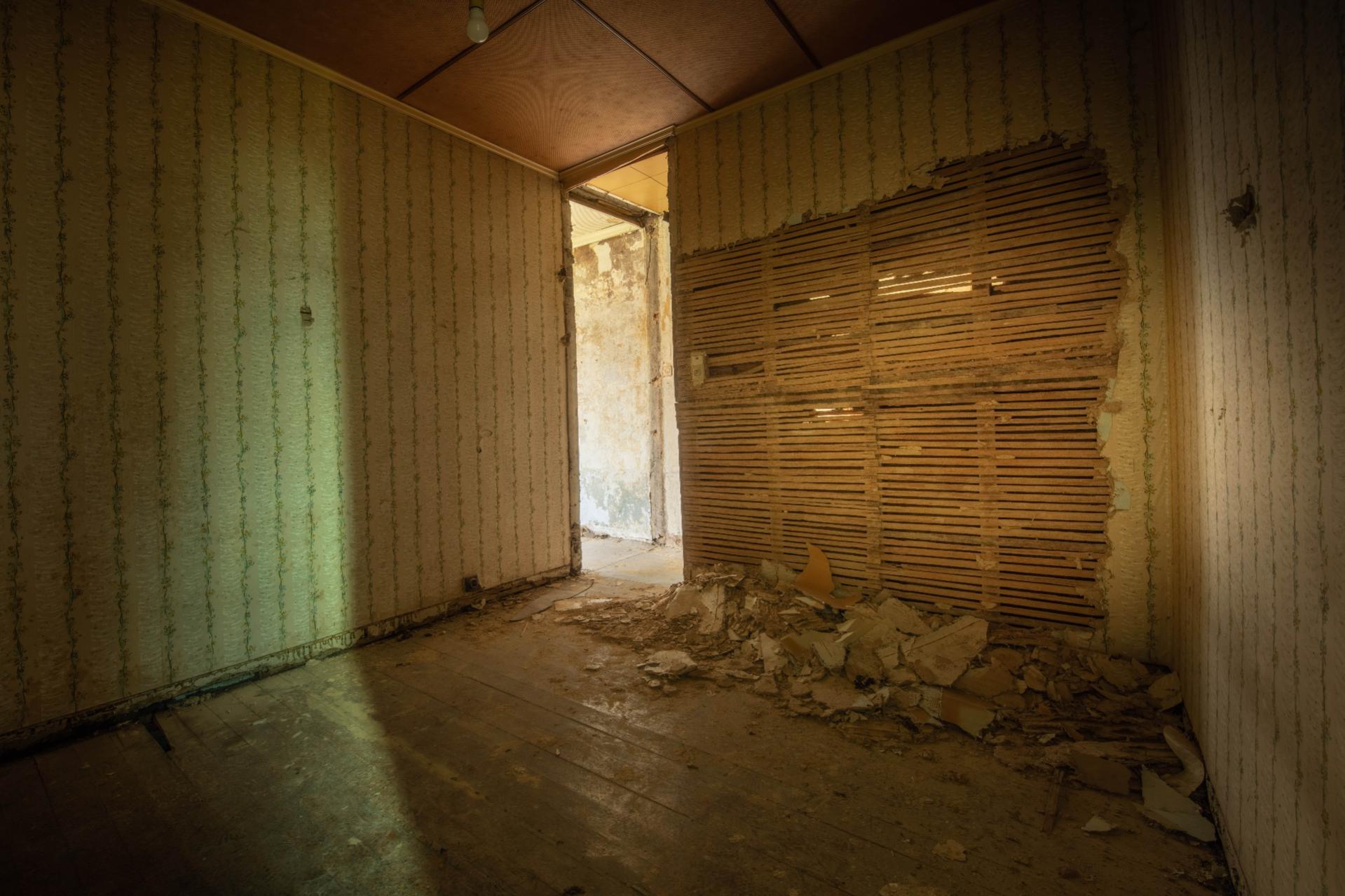 Verlassene Zimmer mit beschädigter Wand, abgenutztem Tapeten und Schutt auf dem Boden. Das Licht fällt durch das Fenster
