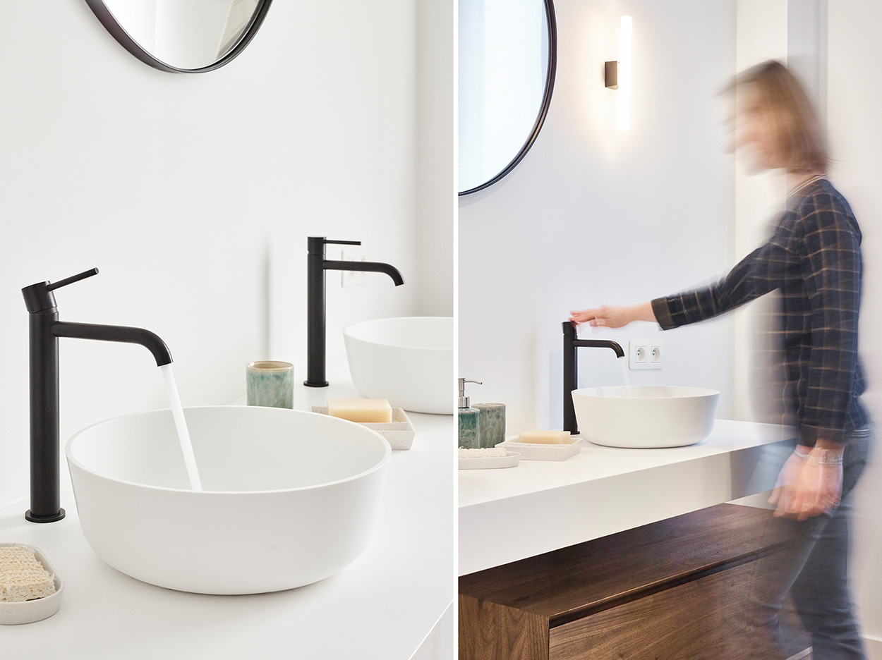 Salle de bain avec des robinets noirs modernes au-dessus d'un lavabo blanc et une armoire en bois, une femme ouvre le robinet.