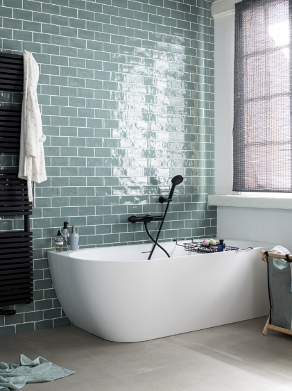 Baignoire autoportante blanche à côté de carreaux verts, radiateur noir et une fenêtre dans la salle de bain.