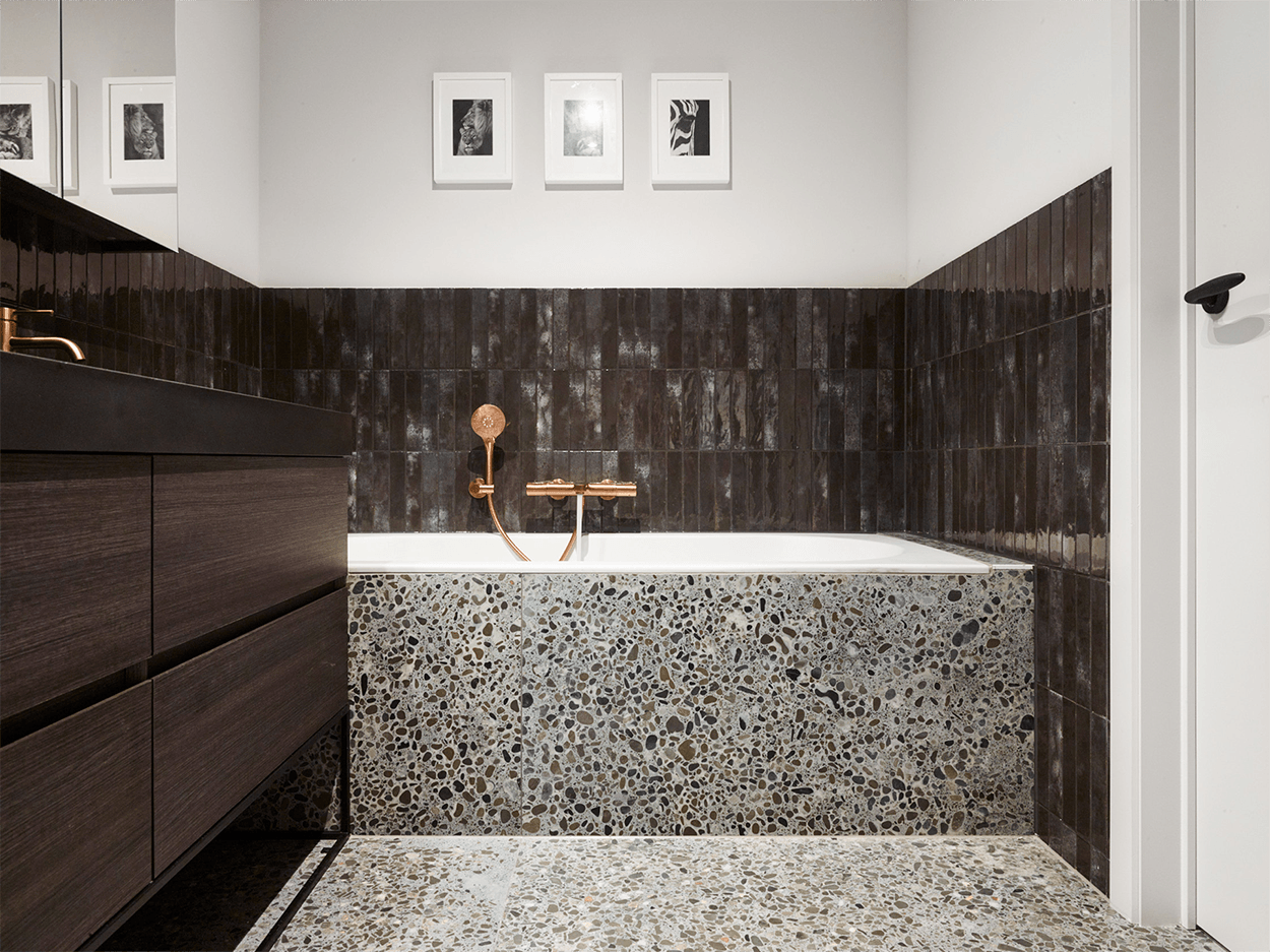 Detail einer Badewanne in einem industriellen Badezimmer von x²o Badezimmer mit Terrazzo-Verkleidung, dunklen Wandfliesen und kupferfarbener Badarmatur.