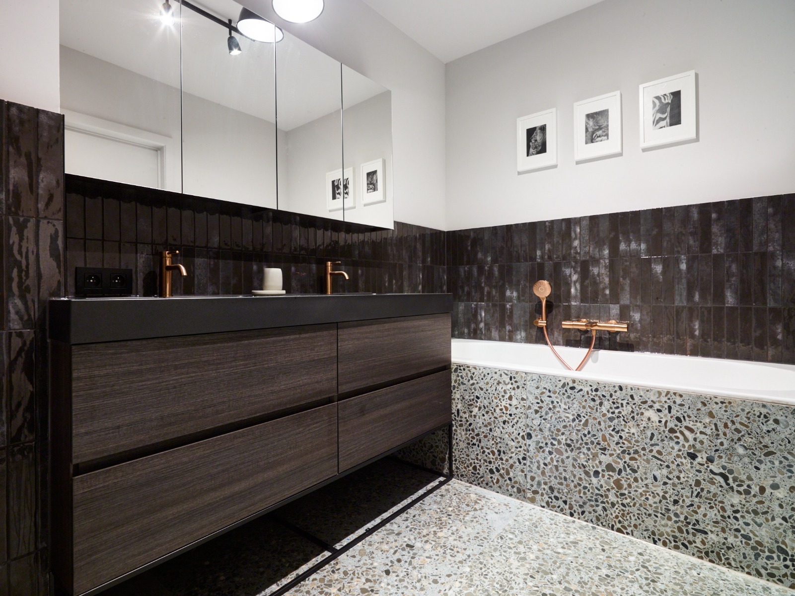 Komplettes industrielles Badezimmer von x²o Badezimmer mit Doppelwaschtisch, Badewanne mit Terrazzo-Verkleidung, Walk-in-Dusche und kupferfarbenen Armaturen in dunkler Farbpalette.