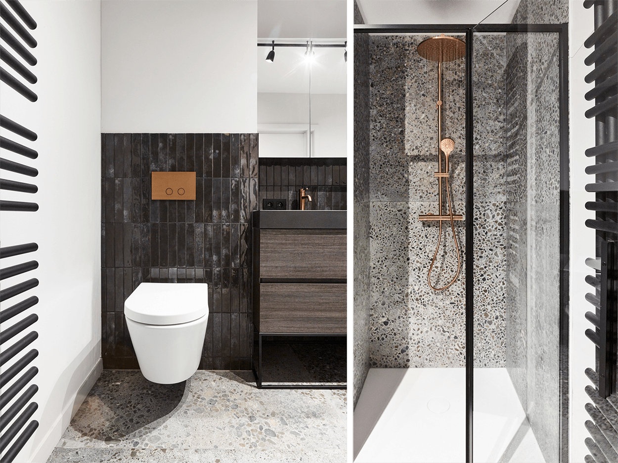 Übersicht eines kompakten industriellen Badezimmers von x²o Badezimmer mit wandhängendem WC, Waschtischunterschrank aus dunklem Holz, Terrazzo-Boden und dunklen Wandfliesen.