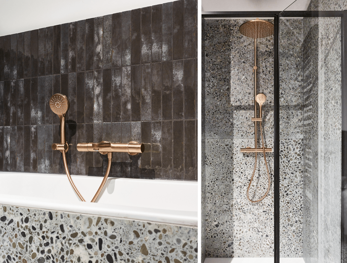 Detailaufnahme einer Badewanne in einem industriellen Badezimmer von x²o Badezimmer mit Terrazzo-Verkleidung, dunklen Wandfliesen und einer kupferfarbenen Badarmatur mit Handbrause.