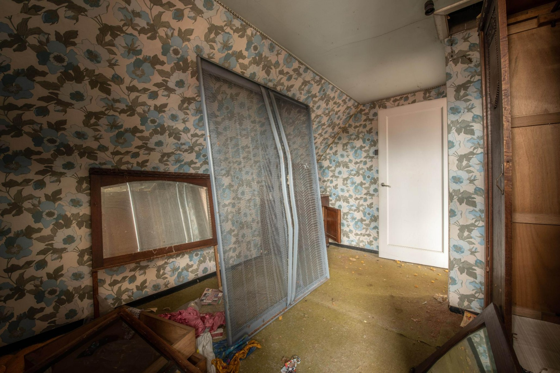 Dieses Vorher-Bild von x²o Badezimmer zeigt einen alten Raum mit auffälliger Blumentapete, einem an die Wand gelehnten Metallbettgestell, losen Spiegeln und beschädigten Möbeln auf einem verschmutzten Boden.