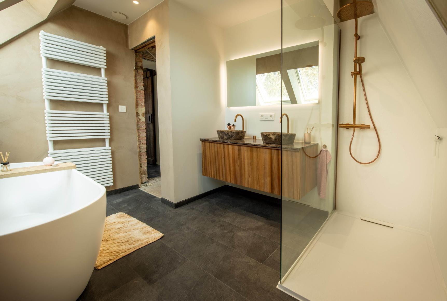 Nach der Renovierung zeigt x²o Badezimmer ein modernes Badezimmer mit freistehender weißer Badewanne, beigen Mikrozementwänden, einer eleganten goldfarbenen Standarmatur, einer Glasduschwand und einem modernen Fliesenboden mit viel Tageslicht durch Dachfenster.